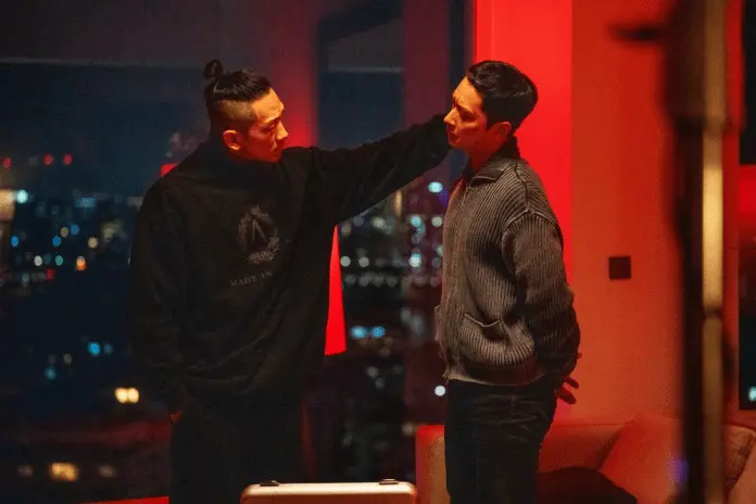 Chansung in a dimly lit room, one with a ponytail wearing a black sweater and the other in a striped gray sweater, engaged in an intense conversation against a backdrop of city lights.