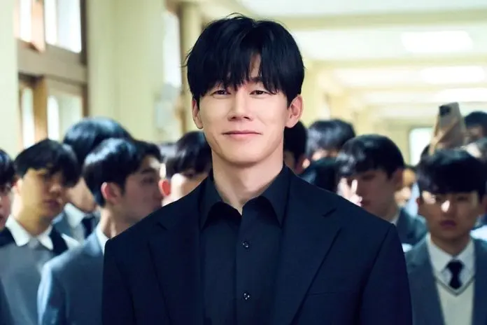 Kim Mu-yeol in a black suit smiles confidently in a school hallway, surrounded by other students in uniform.