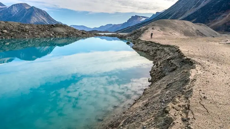 Hiker walking by a lake through remote arctic valley on a partly cloudy summer day. Dramatic arctic landscape of Akshayuk Pass, Baffin Island, Canada. Enormous glacier moraine.