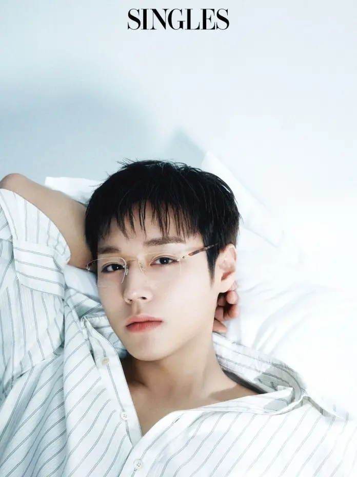 A close-up portrait of a Park Ji-hoon with short hair, wearing glasses and a striped shirt, lying on a white bed. The image has a soft, serene atmosphere.