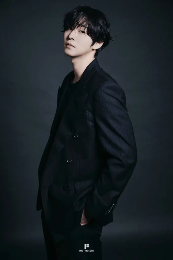 Ahn Hyo-seop in a tailored black suit with pinstripes, standing against a dark background. Their hair is slightly tousled, and they have a contemplative expression.