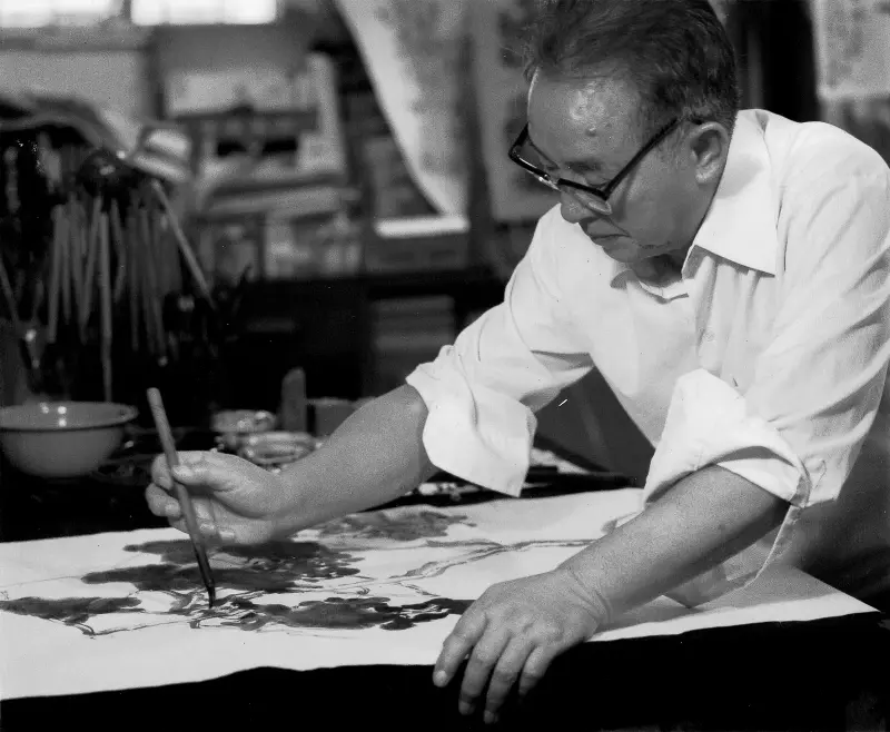 Fan Chang Tien painting with a brush on a large sheet of paper, sleeves rolled up and wearing glasses