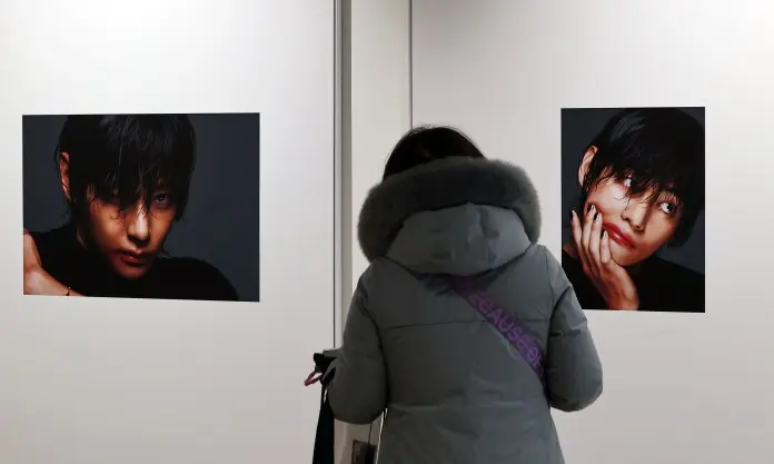 A person in a gray coat is viewing two artistic portraits displayed on a wall, featuring close-up images of V with expressive facial features.