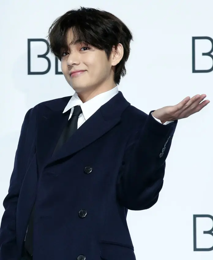V in a dark suit with a white shirt and black tie smiles and gestures with his hand, posing at an event.