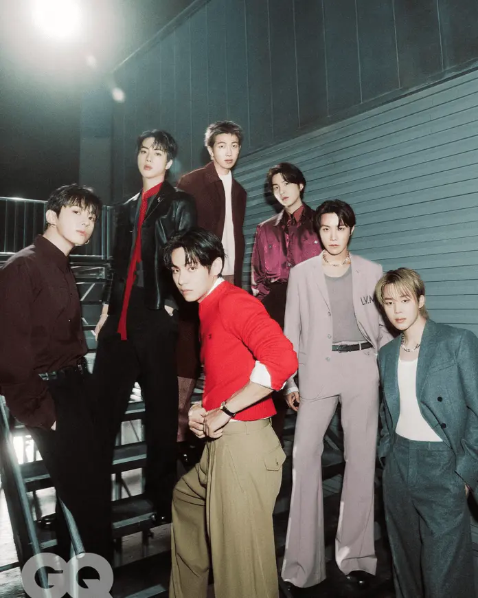 BTS posing together on a staircase, dressed in stylish outfits. The scene is well-lit with a spotlight, highlighting their varied fashion choices.