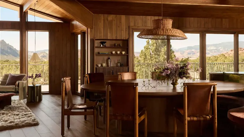 A living-dining area clad in wood with sweeping views of the Utah desert and a large dining table with leather sling seating with wood frames and a large rattan pendant light suspended above it