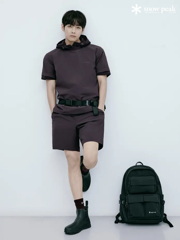 BTS's V dressed in a short-sleeved purple outfit and black rubber boots, leaning against a white wall next to a black backpack.
