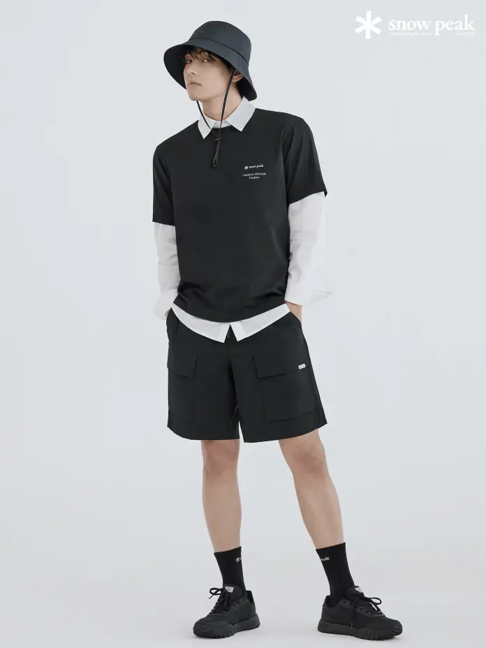 BTS's V dressed in a black t-shirt layered over a white long-sleeve shirt, wearing black shorts and a black bucket hat, standing in a neutral background.