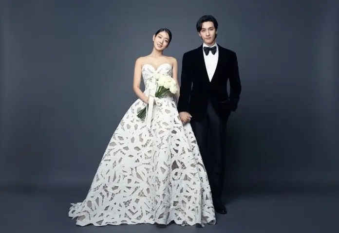 Park Shin-hye and Choi Tae-joon standing together, the woman in a detailed white wedding dress holding a bouquet of flowers, and the man in a formal black tuxedo with a bow tie.