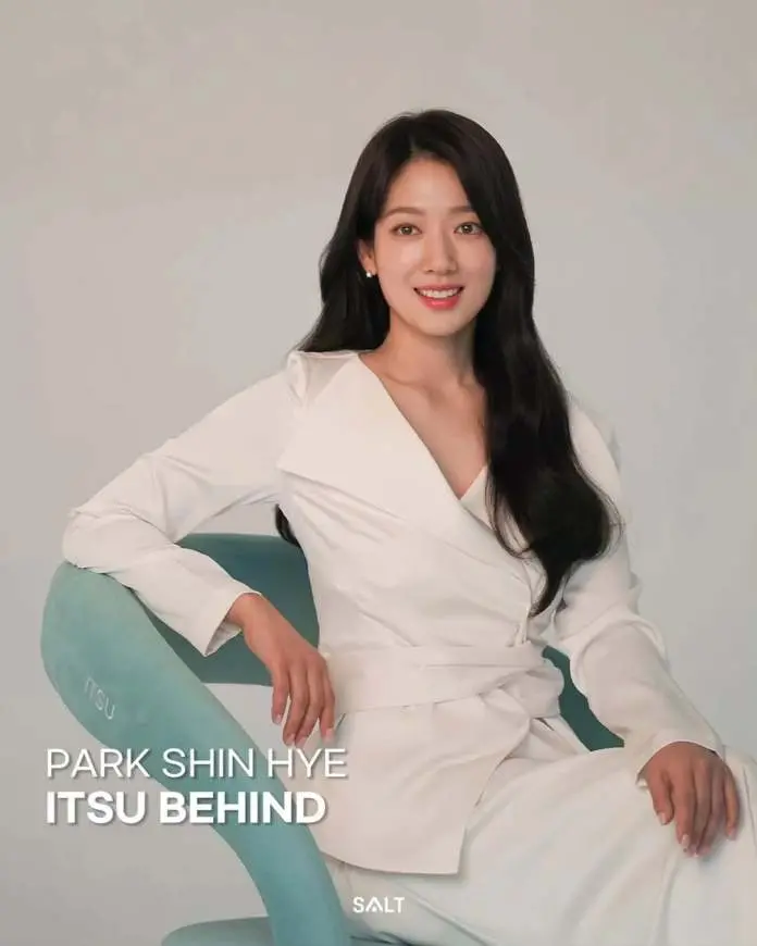 Park Shin-hye in a white outfit sitting on a teal chair, smiling at the camera with a neutral background.