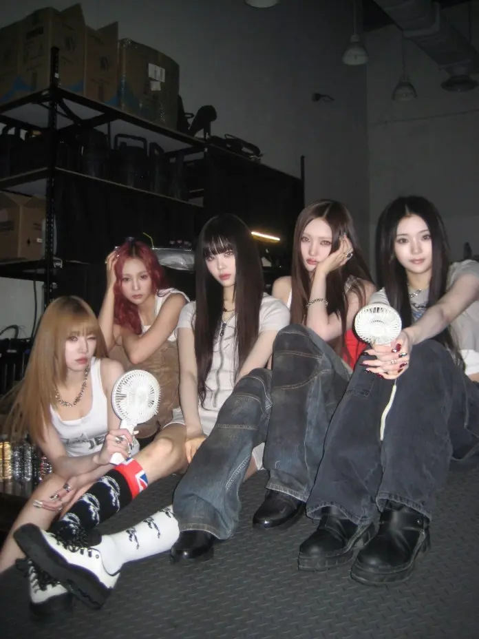 Keyveatz sitting together on a textured surface, dressed in casual outfits, with varying hairstyles. Some are holding small fans, and the background features shelves with boxes.