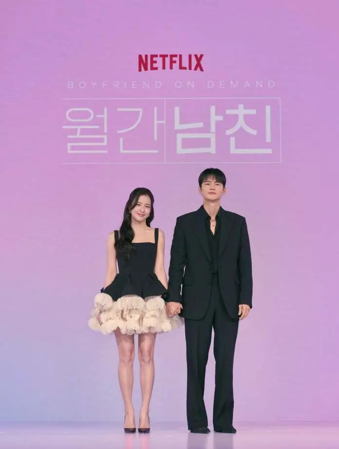 Jisoo and Seo In-guk holding hands at a promotional event for 'Boyfriend on Demand' by Netflix. Jisoo  is wearing a short black dress with a ruffled beige hem, while Seo In-guk is dressed in a black pinstripe suit.