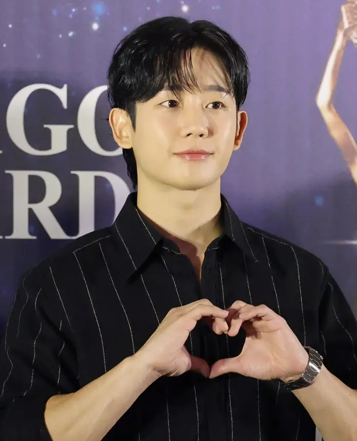 Jung Hae-in with dark hair wearing a black striped shirt forming a heart shape with his hands, smiling at a promotional event.
