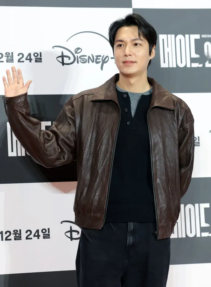 Lee Minho wearing a brown leather jacket and black sweater poses with his hand raised, smiling at a camera, against a backdrop featuring Disney logos.