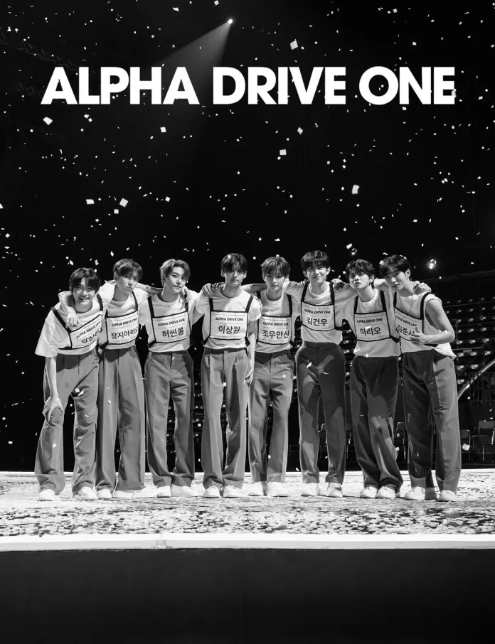 ALPHA DRIVE ONE posing together on stage, wearing matching outfits and name tags, with a stylish logo reading 'ALPHA DRIVE ONE' above them. The background features confetti and a dimly lit arena.