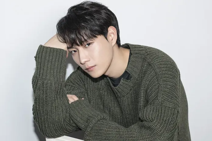 Kim Young Dae with dark hair and a serious expression, wearing a green knitted sweater, leaning on his arm against a light background.