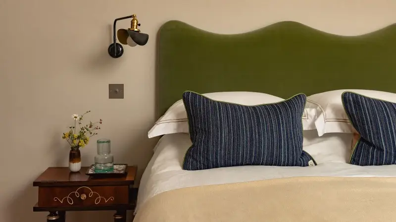 Bedroom with warm neutral walls, a green velvet headboard, striped navy pillows and a dark wood bedside table.
