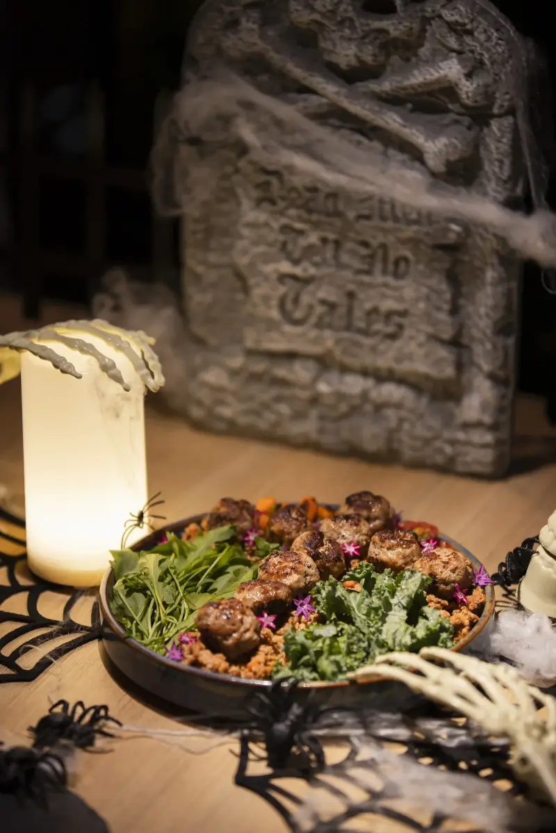 A plated meatball with greens and purple edible flowers, surrounded by spiderwebs, a glowing candle and skeleton-hand props