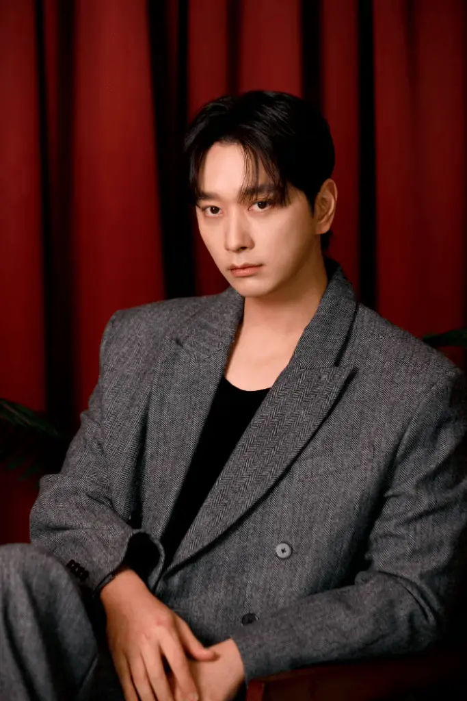 Chansung with dark hair wearing a stylish gray herringbone suit and a black t-shirt, sitting against a red curtain backdrop.
