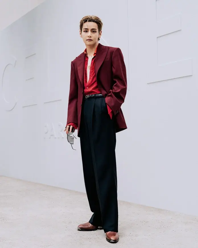 V stands confidently in a stylish outfit featuring a maroon blazer and a red shirt, paired with sleek black trousers and brown shoes. The background showcases a minimalist design with embossed letters.
