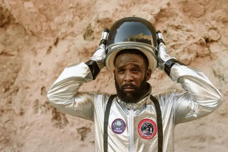 African American astronaut putting on helmet in rocky setting, wearing a shiny silver space suit.