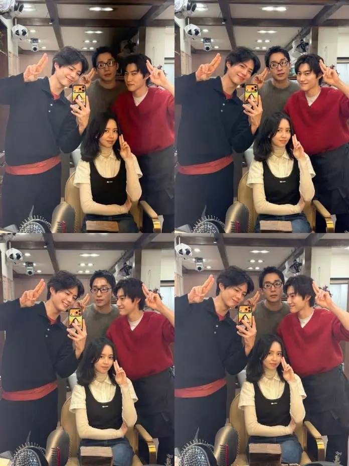A group of four people posing for a selfie in a makeup room, smiling and making peace signs. Two men are wearing sweaters and a third man is wearing glasses, while a woman in a black dress over a white top sits in front.