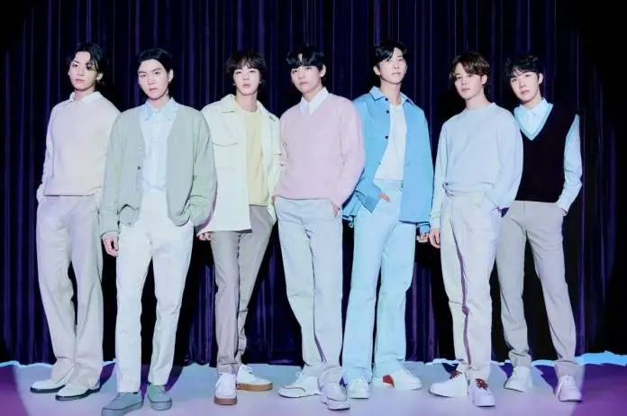 BTS standing in a line against a dark curtain backdrop, dressed in stylish pastel-colored outfits, with each member displaying a unique fashion style.