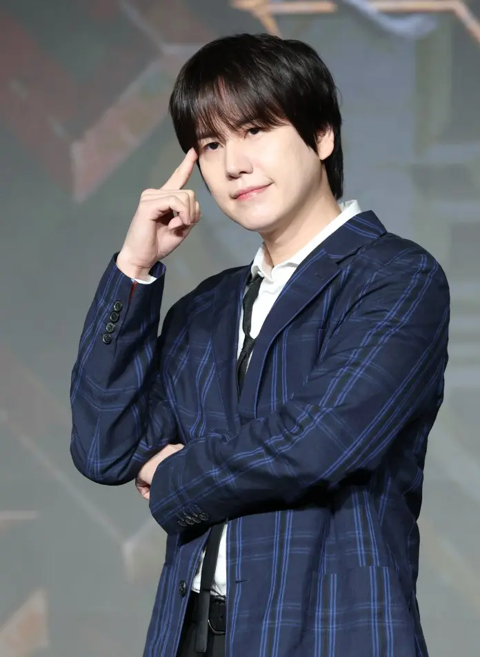 Kyuhyun posing with his hand on his temple, wearing a blue plaid suit jacket over a white shirt and black tie, against a blurred background.