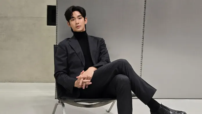 Kim Soo-hyun wearing a black turtleneck and a gray suit sits casually in a modern chair, with a minimalist backdrop.
