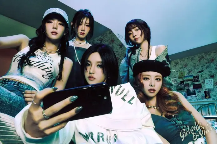 ITZY posing together, looking at a smartphone in a dimly lit room with a floral wallpaper background, wearing casual street-style outfits.