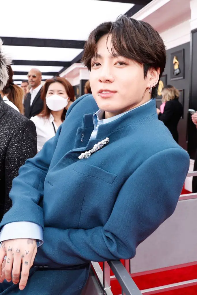 BTS Jungkook in a stylish blue suit, leaning against a railing at a red carpet event, smiling at the camera.
