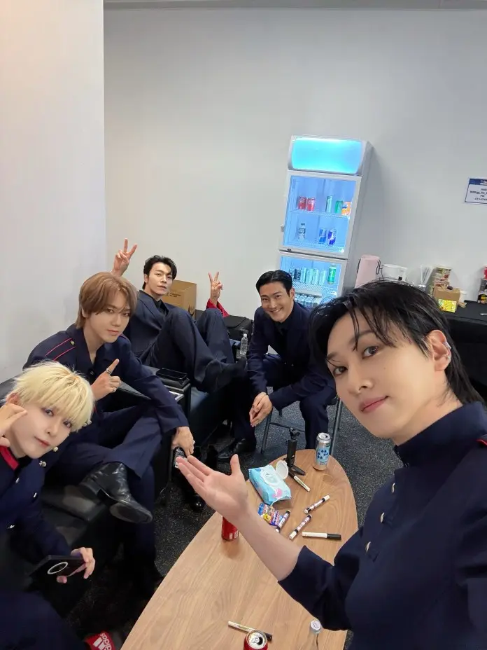 Five individuals in matching navy outfits pose for a selfie in a casual setting, sitting at a table with snacks and drinks. A refrigerator with beverages is visible in the background.