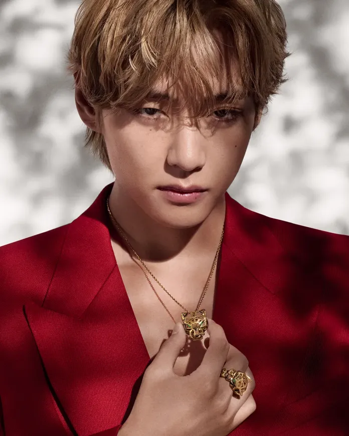 A close-up portrait of a young man with blonde hair, wearing a red suit. He holds a gold pendant and ring featuring a lion design, with a serious expression against a softly blurred background.