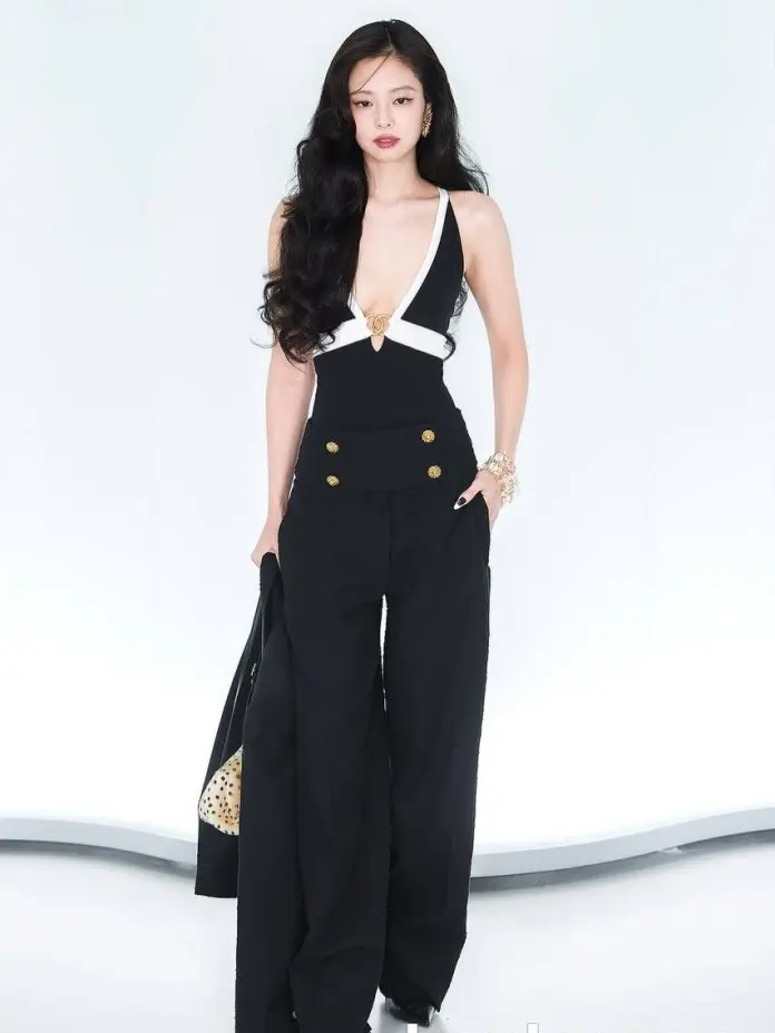 Jennie stands confidently in a stylish black outfit featuring a deep V-neck and wide-leg trousers, accompanied by a handbag. The background is a soft, light color.
