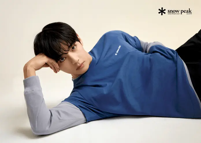 V with dark hair poses casually while lying on a light background, wearing a blue and gray long-sleeve shirt with the logo 'snow peak.'
