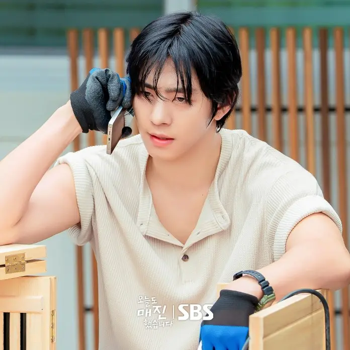 Ahn Hyo-seop with black hair is talking on a smartphone while wearing gloves. He appears focused and is leaning against a wooden structure.