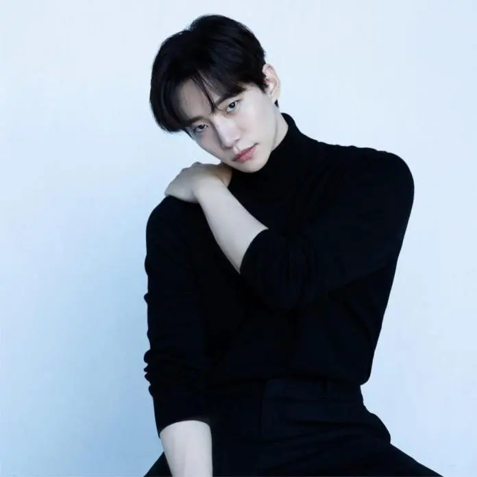 Lee Junho wearing a black turtleneck sits with one arm resting on his shoulder, gazing thoughtfully at the camera against a light blue background.