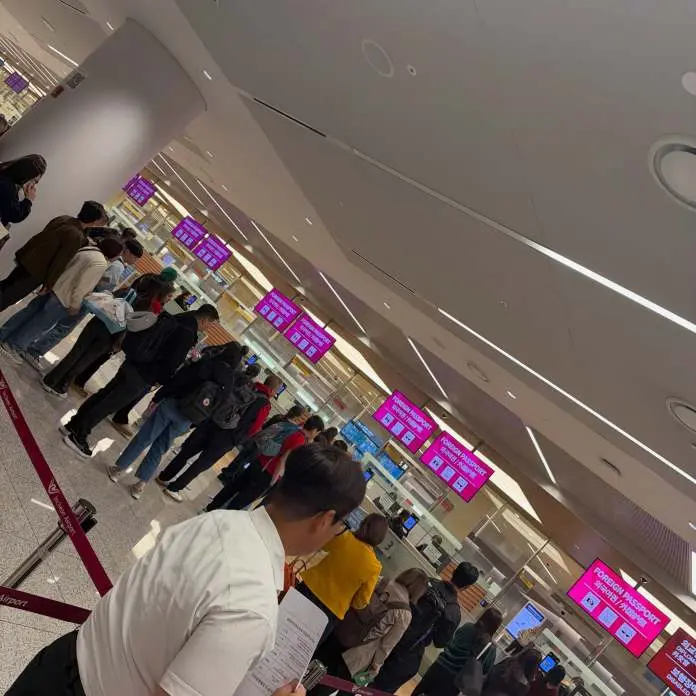 A long line of people waiting at an airport check-in counter, with multiple digital screens displaying information in the background.