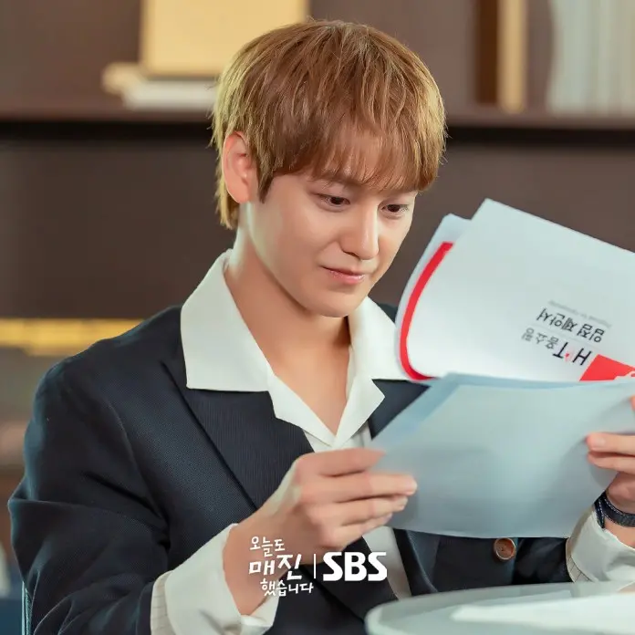 Kim Bum with light brown hair, dressed in a dark suit with a white collar, is sitting at a table and looking thoughtfully at several papers in his hands.