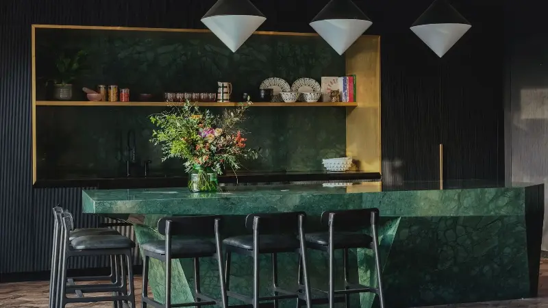 A kitchen island in green marble stone with gold open shelving behind and white pointed pendant lighting above