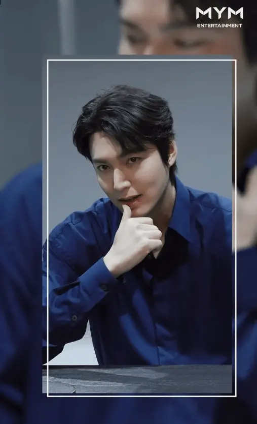 Lee Minho with dark hair poses thoughtfully, resting their chin on their hand while wearing a blue shirt. The background is softly blurred, focusing attention on the individual.