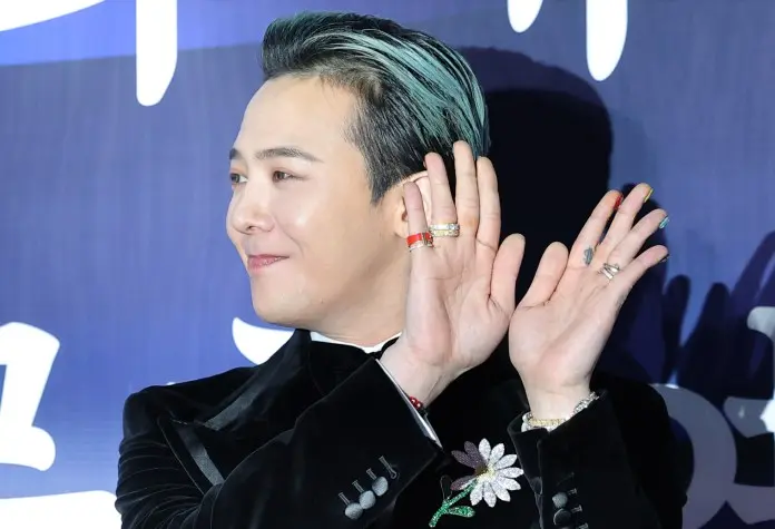 G-Dragon with stylish hair and colorful nail polish raises his hands in a playful gesture, wearing a black velvet suit adorned with decorative rings and a floral accessory.