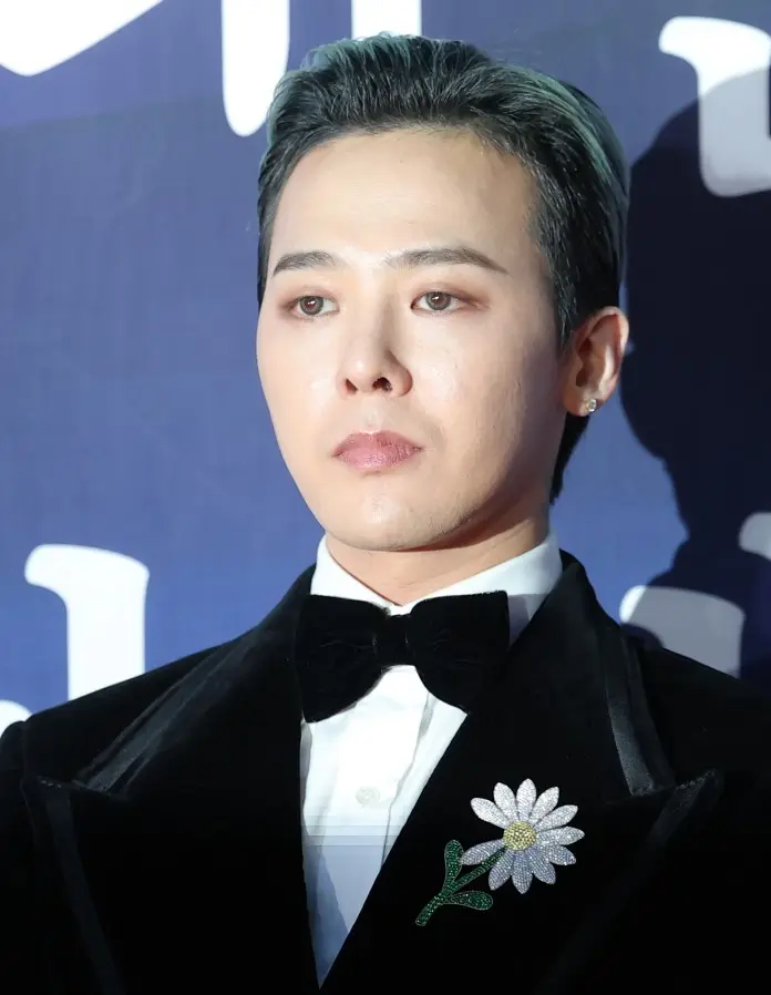 G-Dragon in a black tuxedo with a bow tie, wearing a decorative floral brooch, looking serious against a dark background.