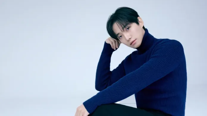 Lee Junho in a navy blue turtleneck sweater sitting thoughtfully against a pale background.