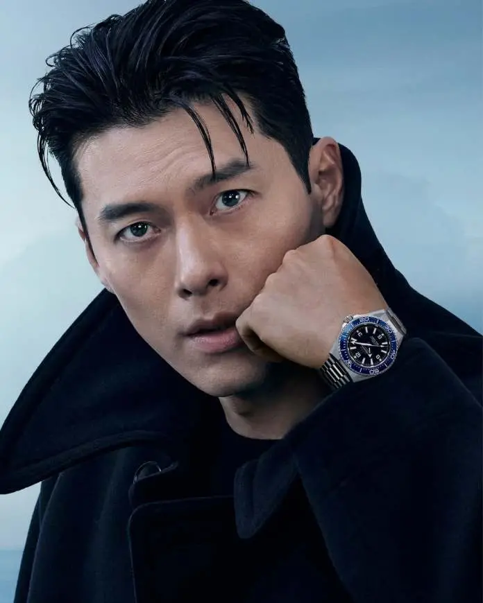 Close-up portrait of Hyun Bin with dark hair styled slicked back, wearing a black coat, resting his chin on his hand while showcasing a silver watch with a blue bezel.