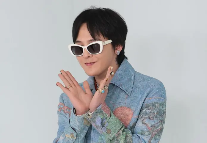 G-Dragon wearing white sunglasses and colorful clothing, striking a playful pose with their hands together. The background is light-colored.