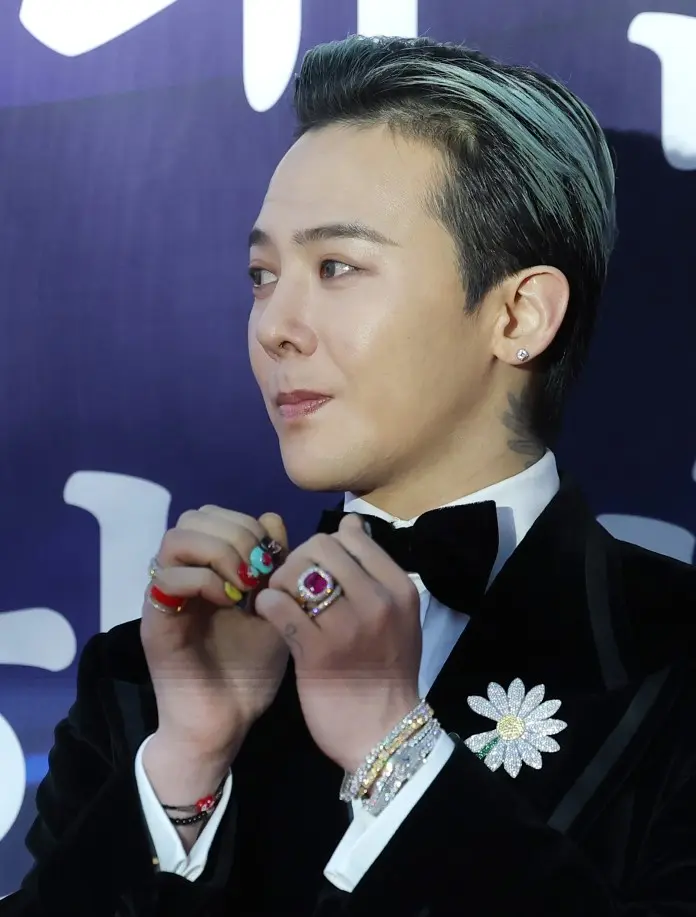 G-Dragon with styled hair, wearing a tuxedo with a bow tie and a decorative flower pin. He is showing colorful nail art on his hands adorned with multiple rings.